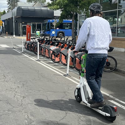 A man rides an e-scooter past a dock full of bike-share bikes. A bus passes through the intersection in the background.